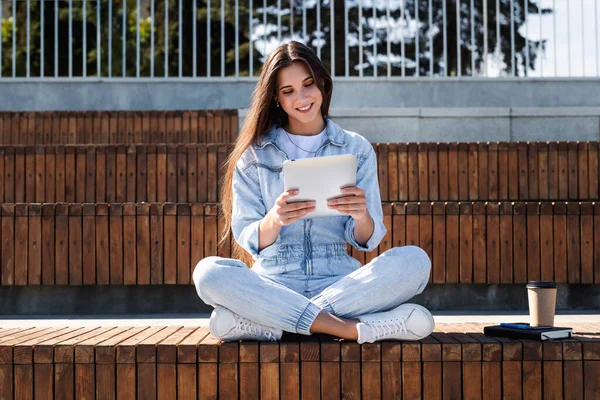 Дівчина тисячоліття в джинсовому одязі сидить на лавці парку, тримаючи в руках розумний планшет. Жінка використовує планшет — стокове фото