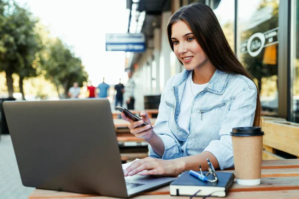 En ung forretningskvinne som jobber på åpen terrasse hjemme, sitter foran en bærbar datamaskin og holder smarttelefon i hånden. – stockfoto