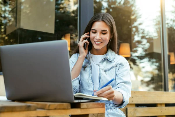 Молода бізнес-леді, що працює в кафе на терасі, сидить перед ноутбуком, розмовляючи по телефону . — стокове фото