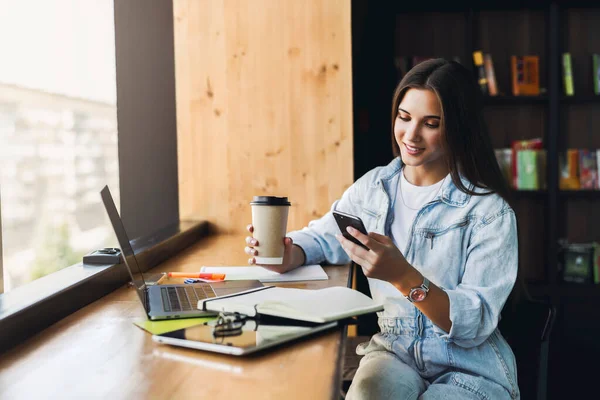 Atractiva mujer de negocios se sienta en la mesa delante de la computadora portátil y habla por teléfono móvil, negocia por teléfono. — Foto de Stock