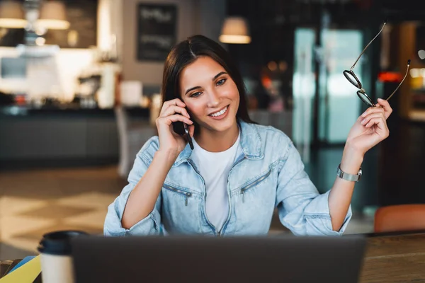 魅力的なビジネス女性はノートパソコンの前のテーブルに座って、携帯電話で話し合い、電話で交渉する. — ストック写真