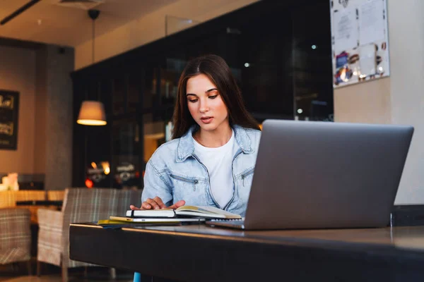 Business woman sits at table in cafe in front of laptop, reads notes from notebook, makes notes in diary.