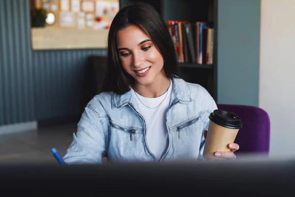 Крупним планом. Красива усміхнена молода бізнес-леді сидить перед ноутбуком, тримає чашку кави, робить нотатки в блокноті — стокове фото