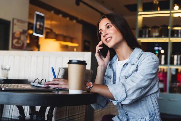Atractiva mujer de negocios se sienta en la mesa delante de la computadora portátil y habla por teléfono móvil, negocia por teléfono. — Foto de Stock