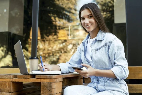 Молода ділова жінка працює на відкритій терасі вдома, сидячи перед ноутбуком, тримаючи в руках смартфон . — стокове фото
