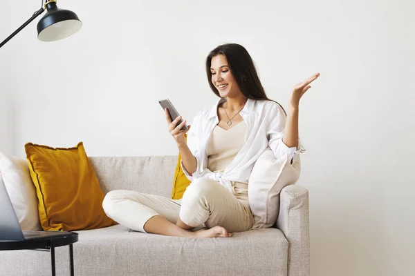 Sorridente bella donna in camicia bianca è seduto sul divano bianco, parlando su smartphone. Donna millenaria — Foto Stock