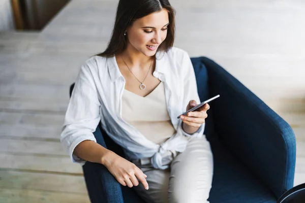 Femme d'affaires souriante assise dans un fauteuil sur fond de mur en bois dans le grenier. Belle fille utilise téléphone mobile — Photo