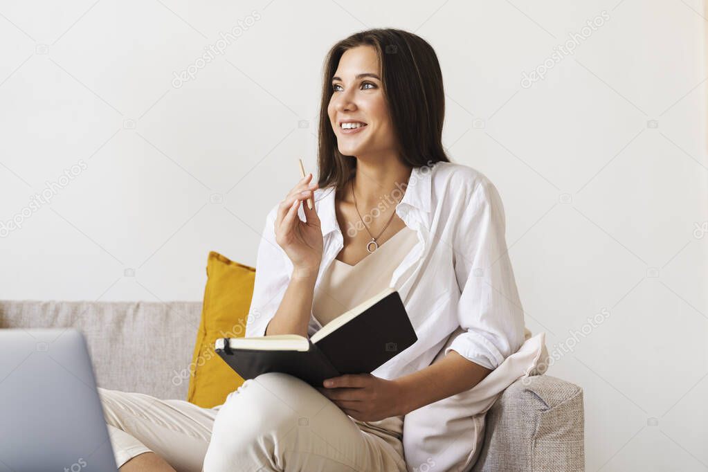Beautiful brunette woman in white shirt sits on sofa in home office, holds pencil, black notepad in her hand. 