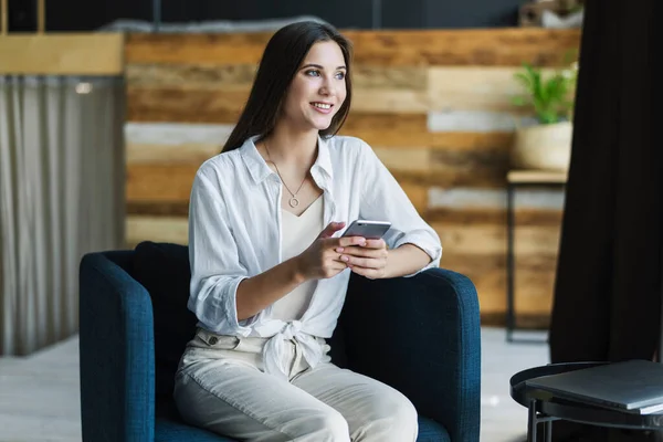 Uśmiechnięta bizneswoman siedząca w fotelu na tle drewnianej ściany na poddaszu. Piękna dziewczyna używa telefonu komórkowego — Zdjęcie stockowe