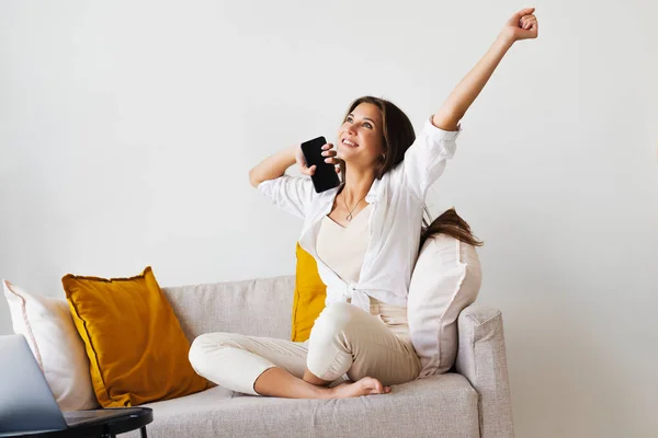 Smiling beautiful woman in white shirt is sitting on white sofa, talking on smartphone. Millennial woman — Stock Photo, Image