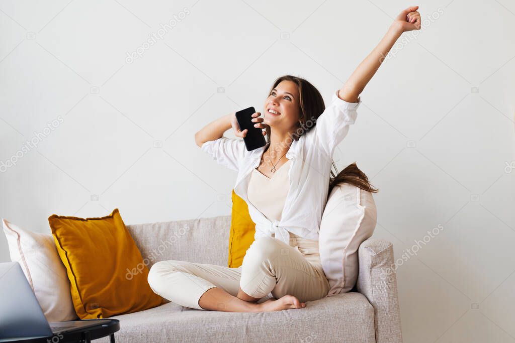 Smiling beautiful woman in white shirt is sitting on white sofa, talking on smartphone. Millennial woman