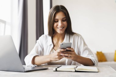 Çekici bir iş kadını laptopun önünde masaya oturur ve iş için akıllı telefonu kullanır..