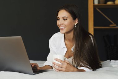 Milenyum kızı elinde bir fincan kahveyle beyaz gömleğiyle yatakta oturuyor. Güzel esmer kadın dizüstü bilgisayar kullanıyor.