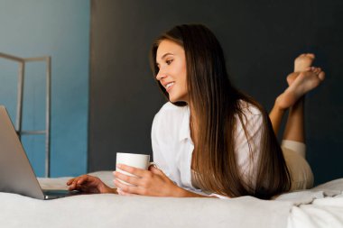 Milenyum kızı elinde bir fincan kahveyle beyaz gömleğiyle yatakta oturuyor. Güzel esmer kadın dizüstü bilgisayar kullanıyor.