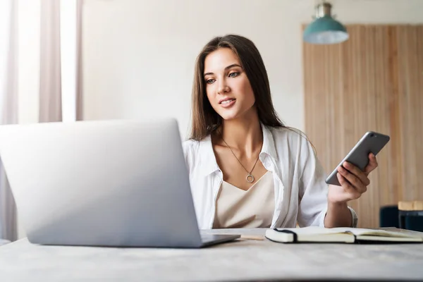 Aantrekkelijke zakenvrouw zit aan tafel voor laptop en maakt gebruik van smartphone voor het werk. — Stockfoto