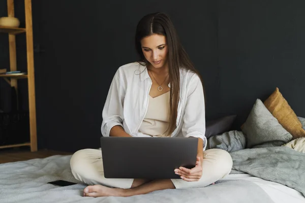 Zakenvrouw gebruikt laptop, werkt op afstand van huis. Glimlachend meisje in wit shirt zit op bed, naast laptop. — Stockfoto