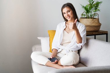 Evde dinlenen bir kadın. Genç milenyum kadını akıllı telefon kullanarak mutlu bir el hareketi yapıyor..