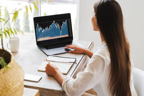 Aantrekkelijke zakenvrouw zit aan het bureau voor laptop en analyseert de aandelenmarkt vanuit haar kantoor. — Stockfoto