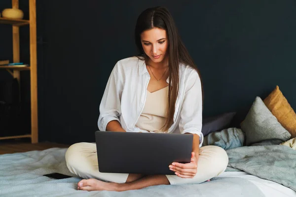 Zakenvrouw gebruikt laptop, werkt op afstand van huis. Glimlachend meisje in wit shirt zit op bed, naast laptop. — Stockfoto