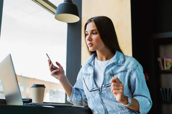 Attraktiv ung kvinne står ved vinduet, holder smarttelefon i hånden, briller for syn og arbeider på laptop. – stockfoto