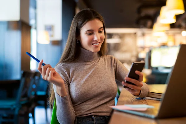 Çekici bir iş kadını laptopun önünde bir masaya oturur ve elinde bir akıllı telefon ve bir kalem tutar.. 