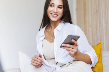 Beyaz gömlekli, gülümseyen sarışın beyaz koltukta oturuyor, elinde akıllı telefonu tutuyor..