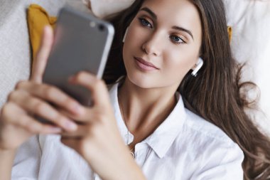 Kadın kanepede uzanıyor, rahatlıyor, akıllı telefonu mesajlar için kullanıyor, fotoğraf değiş tokuşu yapıyor, video izliyor..