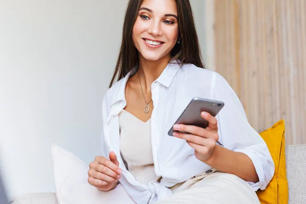 Sorridente bella bionda in camicia bianca siede sul divano bianco, tiene lo smartphone in mano. — Foto Stock