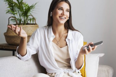 Millennial kızı elinde akıllı telefonuyla beyaz gömleğiyle koltukta oturuyor..