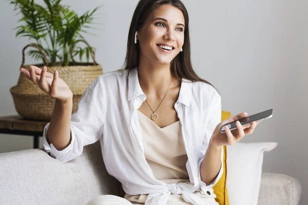 Millennial ragazza è seduta sul divano in camicia bianca, tenendo smartphone in mano. — Foto Stock