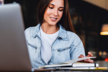 Güzel gülümseyen genç iş kadını dizüstü bilgisayarın önünde oturur, defterine notlar alır..