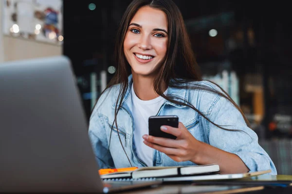 Vrouw lachend, kijkend naar de camera. Aantrekkelijke zakenvrouw zit aan tafel in de voorkant van laptop gesprekken op mobiele telefoon — Stockfoto