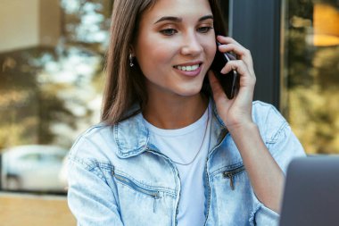 Evde açık terasta çalışan genç bir iş kadını dizüstü bilgisayarın önünde elinde akıllı bir telefonla oturuyor.. 