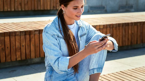 Student siedzi w parku publicznym i korzysta z telefonu komórkowego. Piękna brunetka pisze z przyjaciółmi — Zdjęcie stockowe