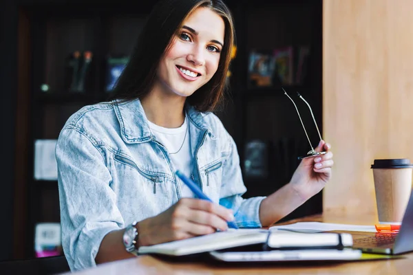 Attraktiv forretningskvinne sitter ved bordet foran laptop 'en. Vakker brunette-kvinne smiler, har briller – stockfoto