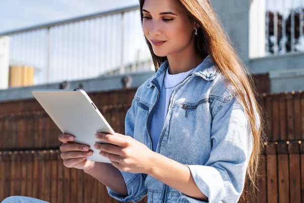 Atrakcyjna kobieta w dżinsowych ciuchach siedzi na ławce w parku, trzyma w rękach inteligentny tablet. Portret pięknej kobiety — Zdjęcie stockowe