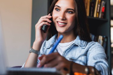 Çekici kadın dizüstü bilgisayarın önünde oturuyor ve cep telefonuyla konuşuyor, not defterine not alıyor.. 