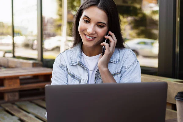 Приваблива ділова жінка, що працює в кафе на відкритій терасі, сидить перед ноутбуком, розмовляє по телефону . — стокове фото
