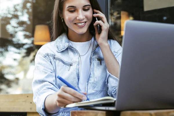 Молода бізнес-леді, що працює в кафе на терасі, сидить перед ноутбуком, розмовляючи по телефону . — стокове фото