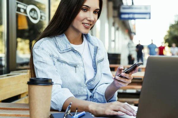 Młoda kobieta biznesu pracująca na otwartym tarasie w domu, siedząca przed laptopem, trzymająca smartfona w ręce. — Zdjęcie stockowe