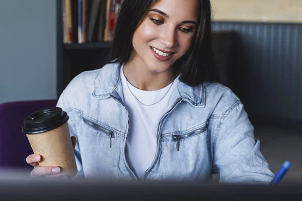 接近中だ。美しい笑顔の若いビジネス女性がノートパソコンの前に座って、コーヒーのカップを保持し、ノートブックにノートを作る — ストック写真