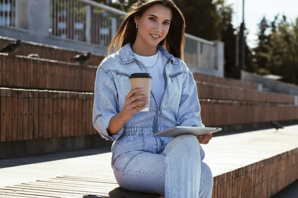 Міленіумна жінка в джинсовому одязі сидить на лавці парку, тримаючи в руках розумний планшет. Жінка використовує планшет Стокове Зображення