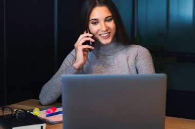Çekici iş kadını dizüstü bilgisayarın önünde oturuyor ve cep telefonuyla konuşuyor.