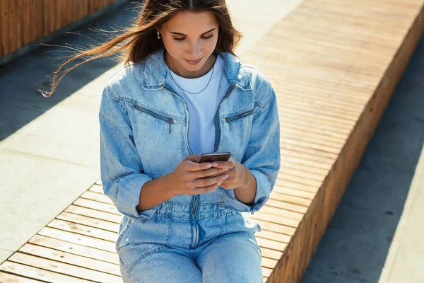 Studentka siedzi w parku publicznym i korzysta z telefonu komórkowego. Piękna brunetka pisze z przyjaciółmi — Zdjęcie stockowe