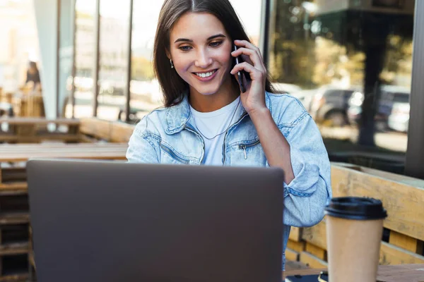 Приваблива ділова жінка, що працює в кафе на відкритій терасі, сидить перед ноутбуком, розмовляє по телефону . — стокове фото