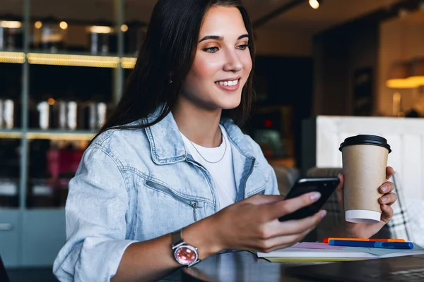 Крупним планом. Красива усміхнена молода бізнес-леді сидить перед ноутбуком, тримає чашку кави, використовує смартфон для роботи — стокове фото