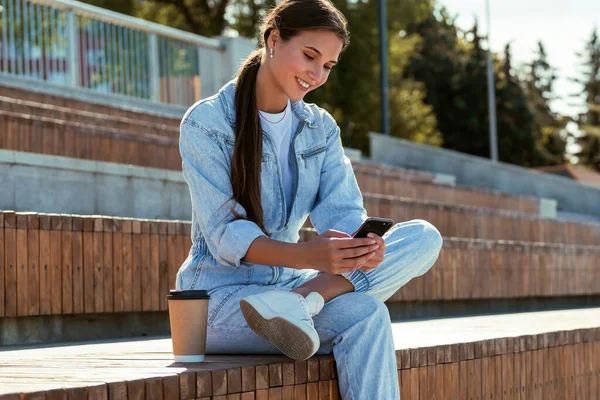 Приваблива дівчина в джинсовому одязі сидить на лавці в парку і пише повідомлення. Красива брюнетка жінка Стокове Зображення