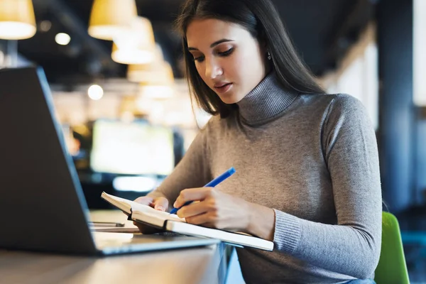 En student forbereder seminar, skriver notater i notatbok, søker informasjon på Internett, bruker Internett, på nettet – stockfoto