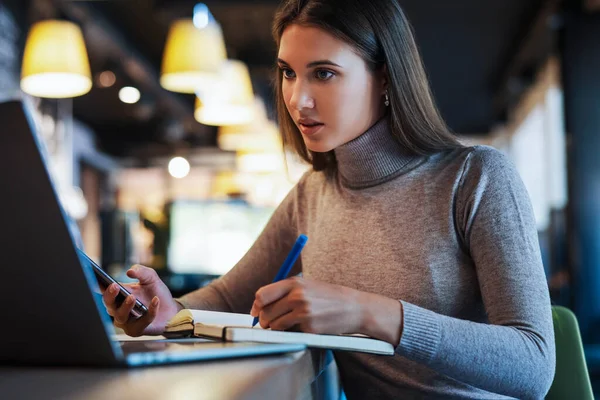 Una mujer hermosa se sienta en una mesa delante de un ordenador portátil y escribe en un cuaderno. — Foto de Stock