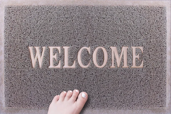 Welcome Door Mat With Female Foot. Friendly Grey Door Mat Closeup with Bare Woman Foot Standing. Welcome Carpet. Girl Foot with White Painted Toenails on Foot Scraper. — Stock Photo, Image